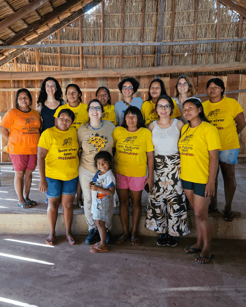 Grupo de 13 mulheres brancas e indígenas em pé dentro de uma estrutura de madeira, as mulheres indígenas estão vestindo camisetas amarelas com os dizeres “um futuro sem incêndios” e “bem viver sem incêndios”. Uma mulher indígena, na ponta esquerda da foto, usa camiseta laranja. Uma criança indígena está sorrindo à frente do grupo.