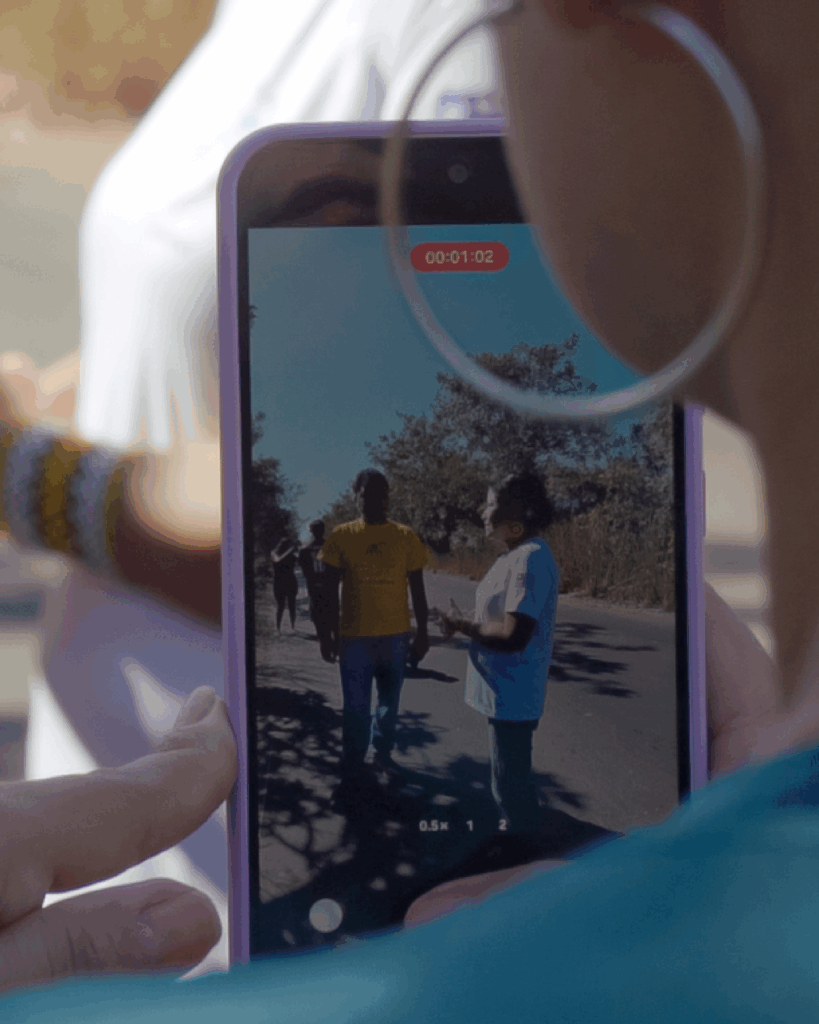 Pessoa usando um brinco de argola segura um celular com capa roxa, que exibe a gravação de um vídeo. Na tela, duas pessoas conversam ao ar livre, com árvores ao fundo — uma veste camiseta amarela e a outra, branca.