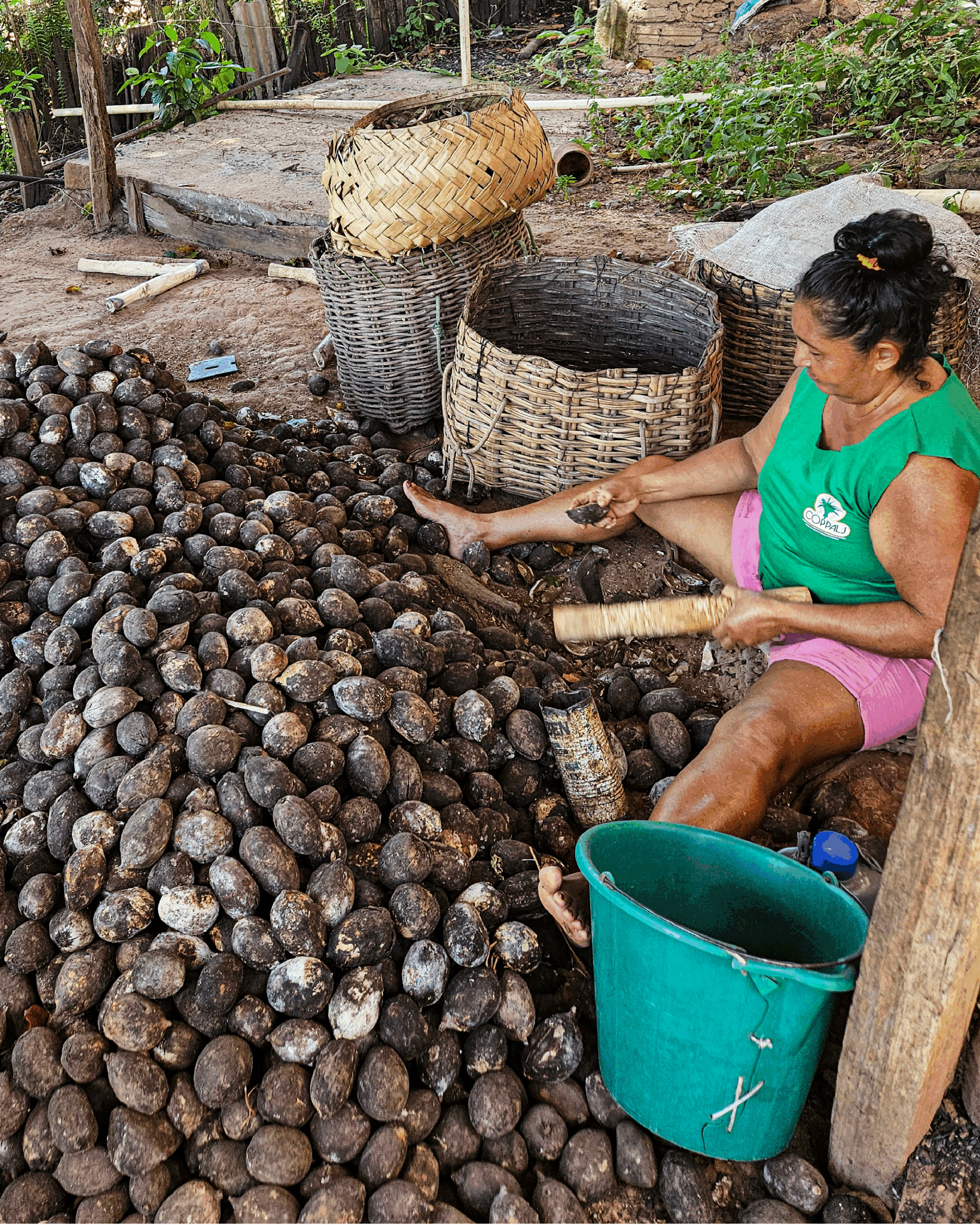Mulher sentada no chão de terra, cercada por uma grande quantidade de sementes escuras de babaçu. Ela segura uma semente em uma mão e uma ferramenta na outra, usada para quebrá-la. Veste camiseta verde e shorts rosa. Ao lado, há três cestos de palha: um vazio e outros dois empilhados um sobre o outro. Ferramentas e objetos estão espalhados ao redor. A cena retrata uma prática tradicional de beneficiamento do babaçu, comum em comunidades agroextrativistas do Cerrado.