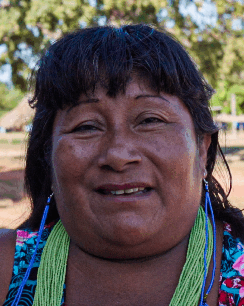 Mulher de cabelos longos e escuros, olhos pretos, pele escura, usando brincos azuis, aparece em primeiro plano, olhando para a câmera. Ao fundo, há árvores e uma estrutura desfocada, sugerindo ambiente externo.