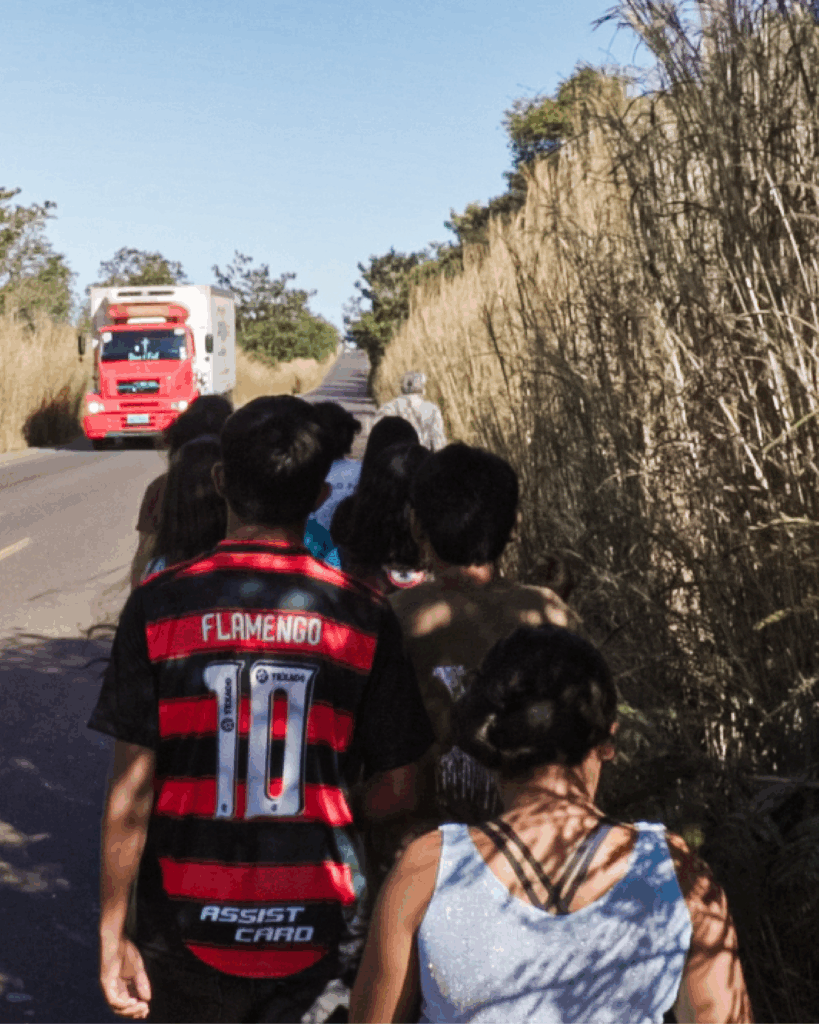 Grupo de pessoas caminha por acostamento ladeado por capim alto à direita. Em primeiro plano, uma pessoa veste camisa listrada vermelha e preta com o nome “Flamengo” e o número 10 nas costas. Ao fundo, um caminhão vermelho se aproxima pelo asfalto.
