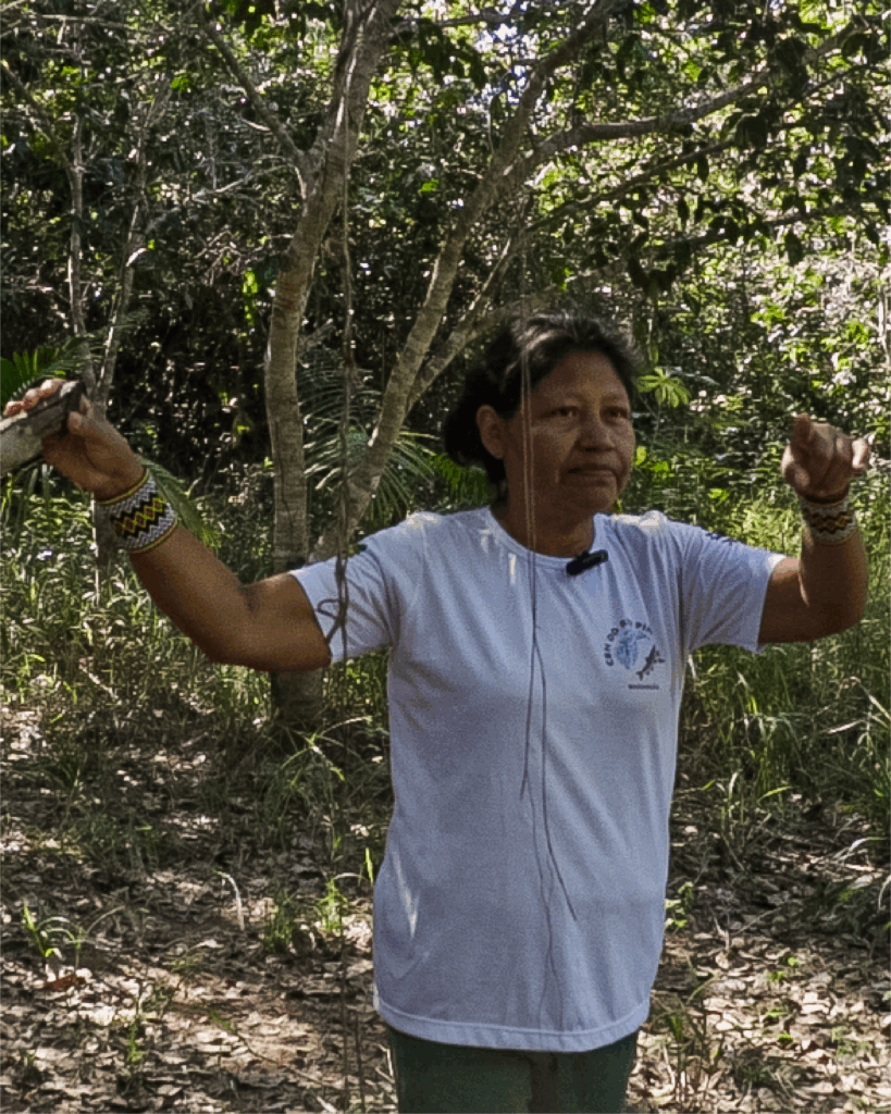 Mulher indígena em pé em área de mata, usando camiseta branca com logotipo no peito. Tem cabelos lisos, escuros e presos. Usa pulseiras coloridas nos dois pulsos. Ao fundo, há árvores e vegetação do Cerrado.