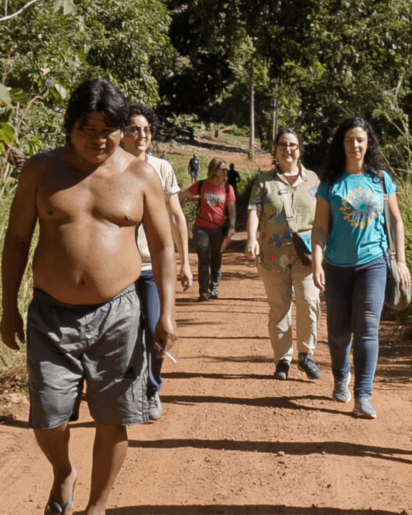 Grupo de 6 pessoas caminha por estrada de terra cercada por vegetação. Em primeiro plano, um homem sem camisa, de bermuda e de chinelo caminha à frente do grupo; os demais usam roupas casuais. A cena ocorre em ambiente de vegetação verde, com árvores ao fundo.