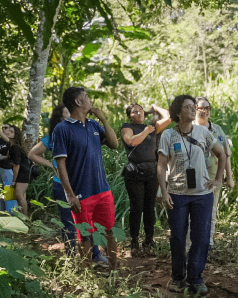 Grupo de 7 pessoas em pé ao ar livre, cercado por vegetação verde e árvores. Algumas usam roupas casuais, como camiseta branca com crachá e camisa polo azul com bermuda vermelha.