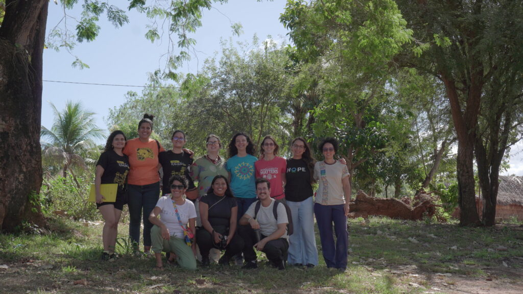 Grupo de onze pessoas posa ao ar livre em meio à vegetação, com árvores e uma palmeira ao fundo. Algumas estão em pé, outras ajoelhadas, vestindo roupas casuais e camisetas com estampas variadas.